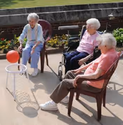 Three elderly women sitting outdoors on a patio, with one woman in a wheelchair and another woman holding a red ball near a small basketball hoop.