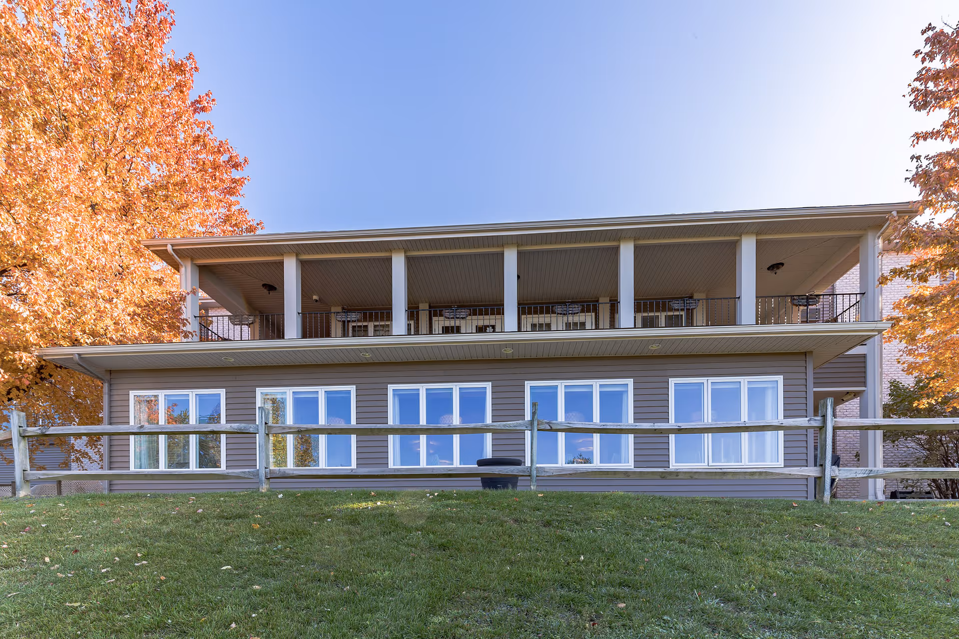 Front exterior of a two-story building with a covered upper balcony, large windows, a wooden fence, and autumn trees.