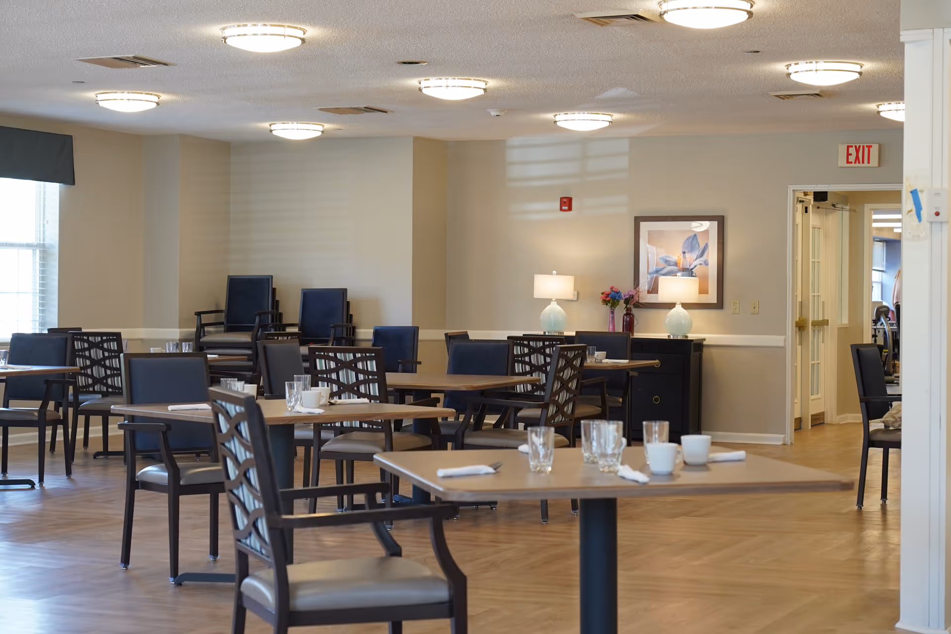 Empty dining room with multiple set tables and chairs, a sideboard with lamps and artwork, and an exit door in the background.