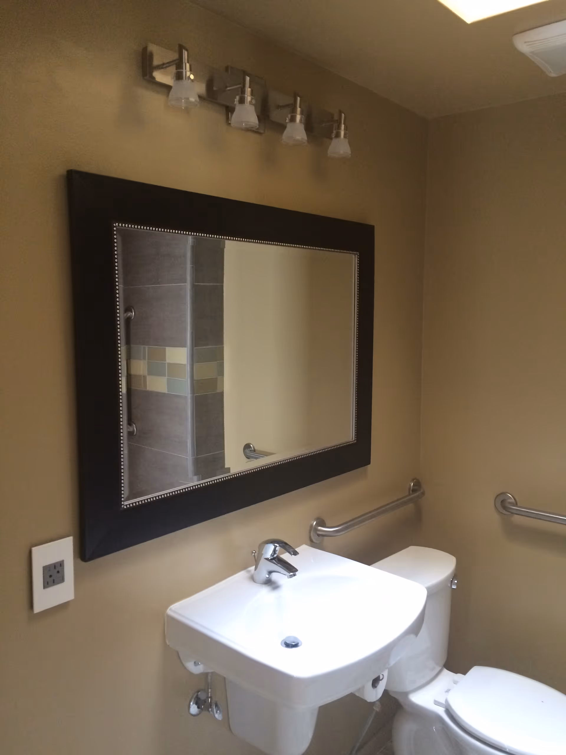 A bathroom with beige walls featuring a white sink with a modern faucet, a white toilet, and a large rectangular mirror with a black frame above the sink. There are metal grab bars on the walls near the toilet and sink, and a light fixture with four bulbs above the mirror. A tiled shower area is partially visible in the mirror's reflection.