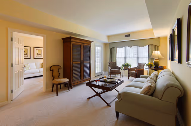 A cozy living room with a light green sofa, a wooden coffee table with a tray and decorative items, two armchairs near a large window with curtains, a wooden armoire, and a side table with a lamp and flowers. An adjacent room with a white sofa and framed artwork is visible through an open doorway.