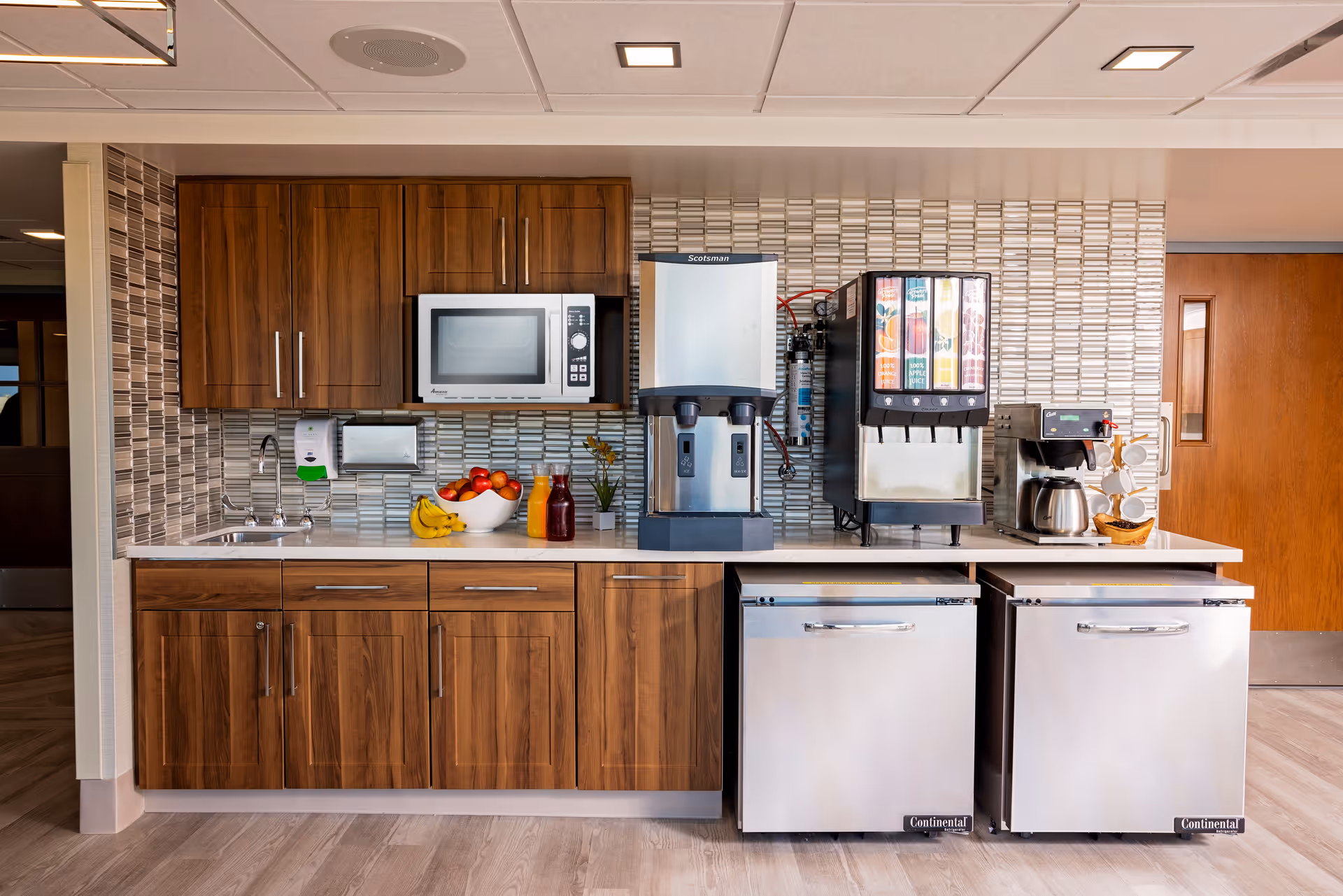 A kitchenette area with wooden cabinets, a microwave, a sink, a bowl of fruit, juice bottles, a coffee machine, a soda dispenser, and two small refrigerators underneath the counter. The backsplash features a modern tile design, and the floor is light wood.