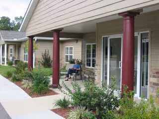 A covered outdoor walkway at Garden Gate Estates with maroon columns, beige siding, and a person sitting on a bench near the entrance of a unit numbered 5230. The area is landscaped with green shrubs and plants along the sidewalk.