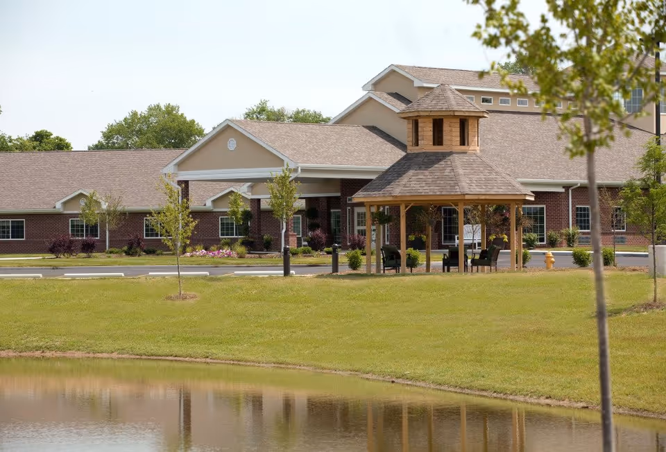 Exterior view of Bridgewater Healthcare Center showing a brick building with a covered entrance and a wooden gazebo surrounded by green grass, small trees, and a pond in the foreground.