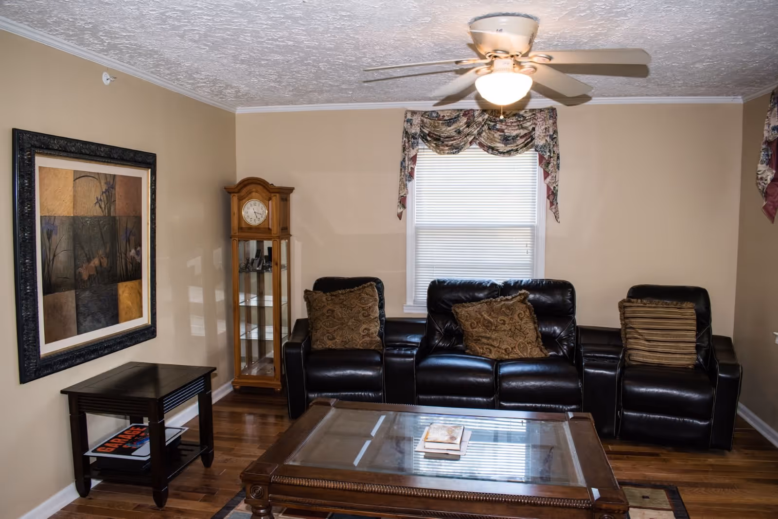 A cozy living room with a black leather sofa and two matching armchairs, each with decorative pillows. A wooden grandfather clock stands in the corner next to a window with floral valance curtains. A glass-top wooden coffee table with books on it is in front of the seating area, and a small dark wooden side table with magazines is positioned against the wall. The room has beige walls, a ceiling fan with a light, and hardwood floors.