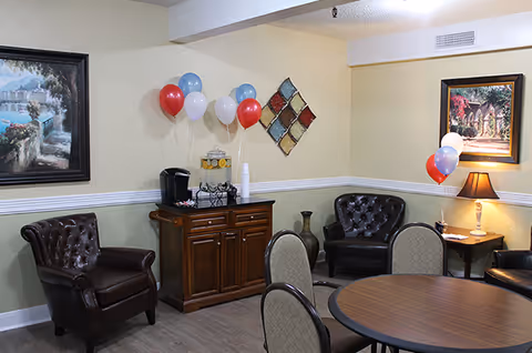 A cozy common area with a round wooden table surrounded by chairs, two dark brown leather armchairs, a wooden cabinet with a beverage dispenser and cups, and decorative balloons in red, white, and blue. The walls are adorned with framed paintings and a colorful diamond-shaped wall decoration. A table lamp is lit on a side table in the corner.