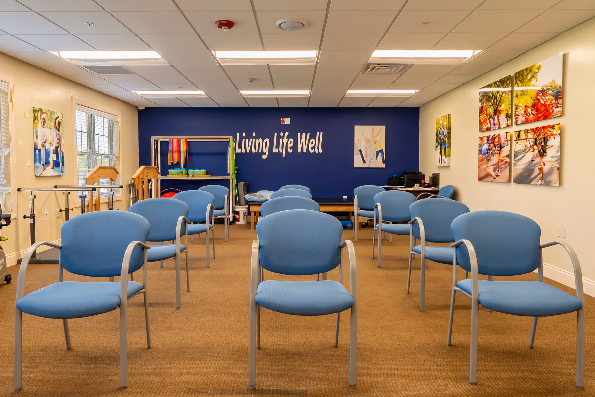 A room with multiple blue chairs arranged in rows facing a blue wall with the words 'Living Life Well' written on it. The room has beige carpet, white ceiling tiles with fluorescent lights, and several colorful pictures on the walls. There are windows on the left side letting in natural light and some exercise or therapy equipment near the back wall.