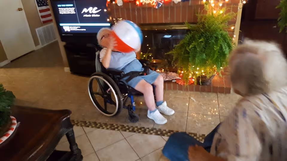 An elderly person in a wheelchair playing with a beach ball indoors near a fireplace decorated with string lights and a large green plant. Another elderly person is seated nearby, partially visible. The room has tiled flooring and a television in the background.