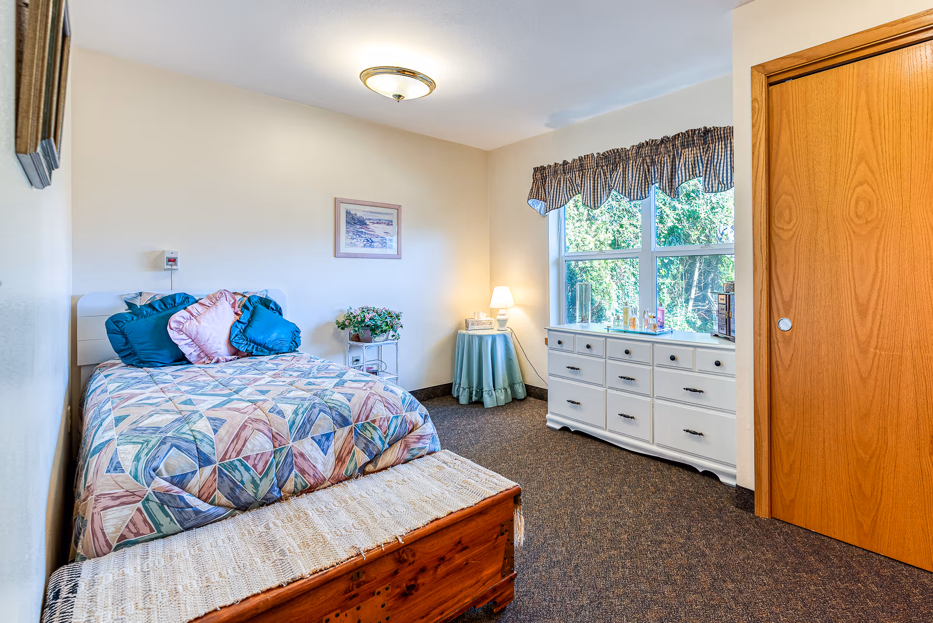 A cozy bedroom in an assisted living facility featuring a bed with colorful geometric patterned bedding and decorative pillows. There is a wooden chest at the foot of the bed, a small round table with a lamp and tissue box, a white dresser with various items on top, a large window with a checkered valance, and a wooden closet door.