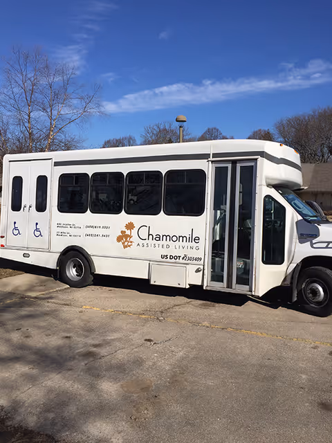 A white shuttle bus parked outdoors on a sunny day with clear blue sky. The bus has the logo and name 'Chamomile Assisted Living' on its side along with contact information and accessibility symbols. Trees without leaves are visible in the background.