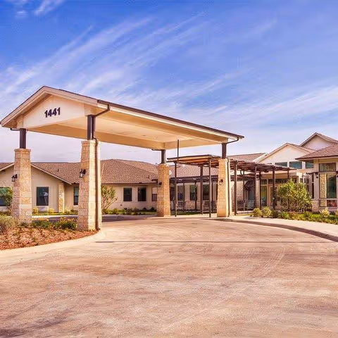 Exterior view of a senior living facility with a covered entrance supported by stone pillars, a driveway, and a clear blue sky with some clouds.