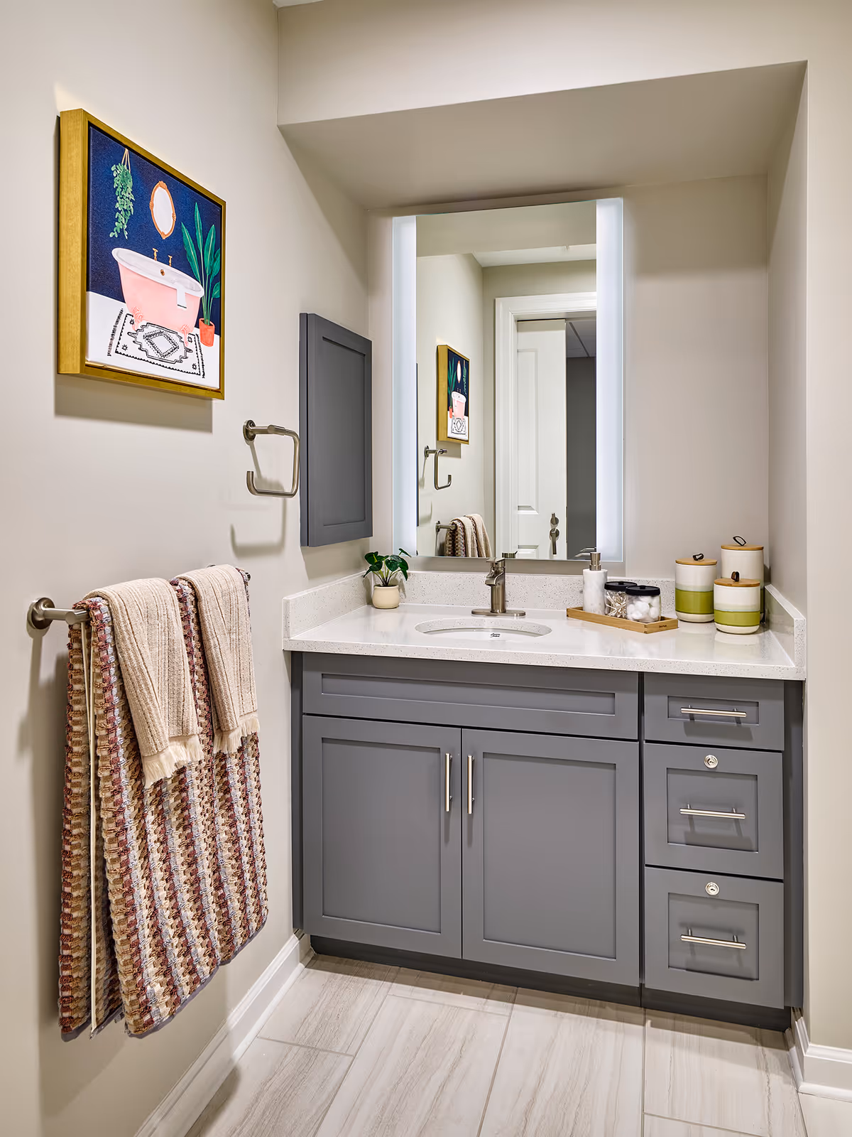 A bathroom vanity area with a gray cabinet, white countertop, and an undermount sink. Above the sink is a large rectangular mirror with built-in lighting on both sides. To the left, there is a towel rack with two towels hanging and a framed artwork of a pink bathtub and plants on the wall. The floor is tiled in light colors.