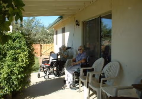 Two elderly individuals sitting outside on a covered patio area of a residential facility. One person is in a wheelchair and the other is seated on a plastic chair. The patio is adjacent to a building with sliding glass doors and there are additional empty chairs nearby. Green bushes and a wooden fence are visible in the background under a clear sky.