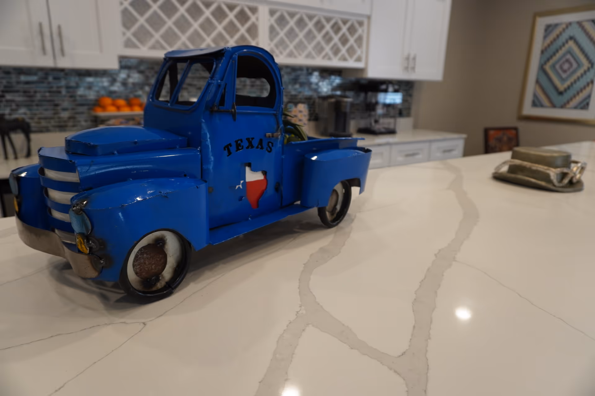 A close-up of a blue decorative metal toy truck with the word TEXAS and a Texas state shape painted on its door, placed on a white marble countertop in a kitchen. The background shows white cabinets, a tiled backsplash, a bowl of oranges, and some kitchen appliances.