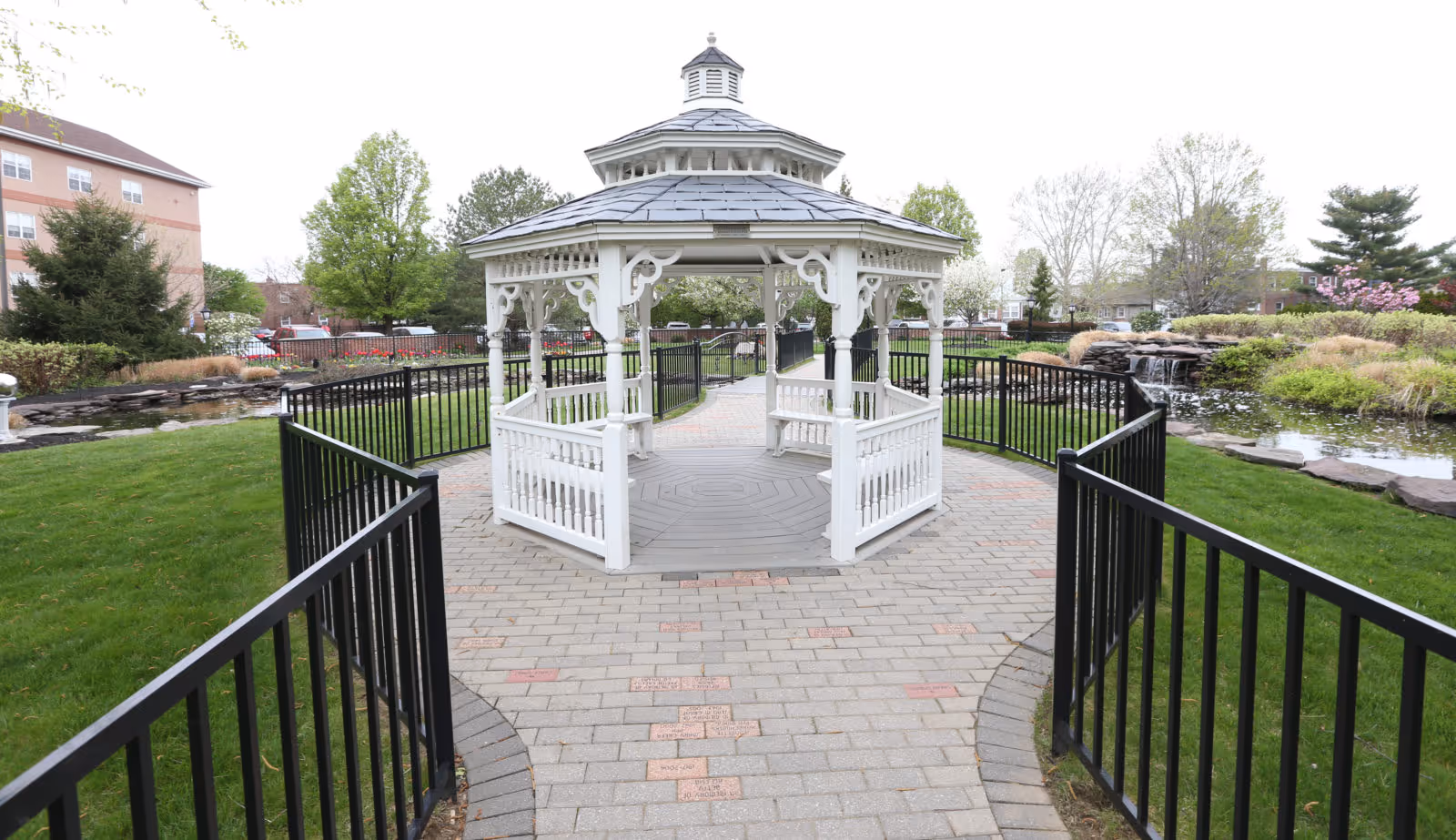 A white wooden gazebo with a gray shingled roof situated on a paved walkway surrounded by black metal railings. The gazebo is located in a landscaped garden area with green grass, trees, bushes, and a small pond with a waterfall feature. A multi-story building is visible in the background.