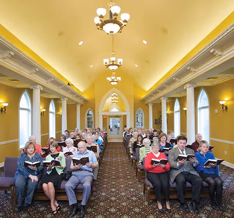 A large group of elderly people seated in rows inside a spacious room with high vaulted ceilings, chandeliers, and arched windows. They are holding and reading from books, possibly hymnals or prayer books, suggesting a communal or religious gathering.