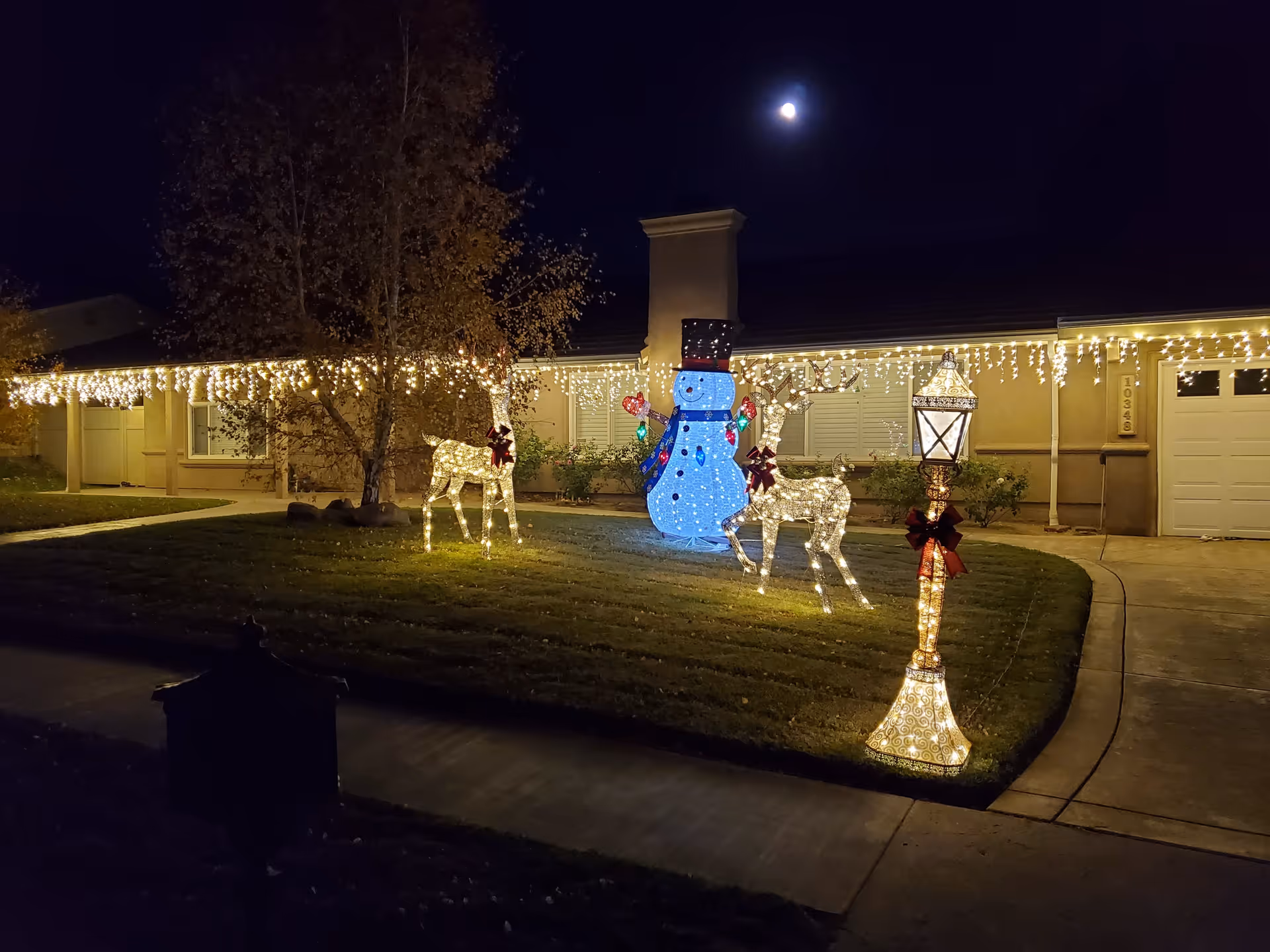 Nighttime view of a house decorated with Christmas lights including icicle lights along the roofline, two illuminated reindeer, a large glowing snowman, and a lit streetlamp with a red bow on the front lawn.