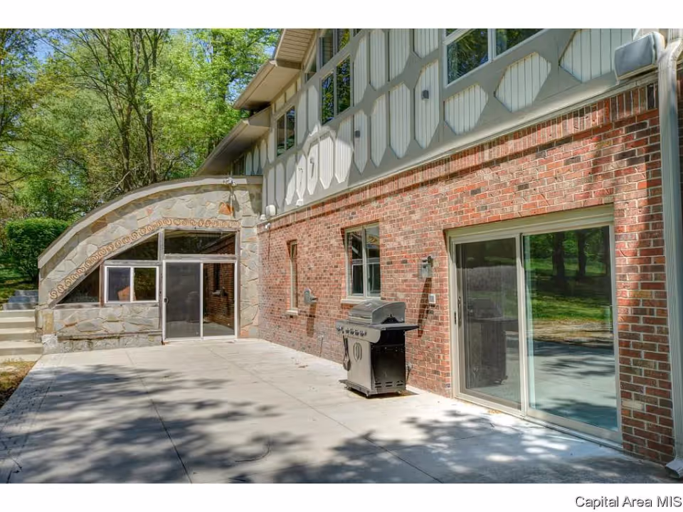 Outdoor patio area adjacent to a brick building with large windows and sliding glass doors. There is a barbecue grill on the concrete patio, and trees and greenery are visible in the background.