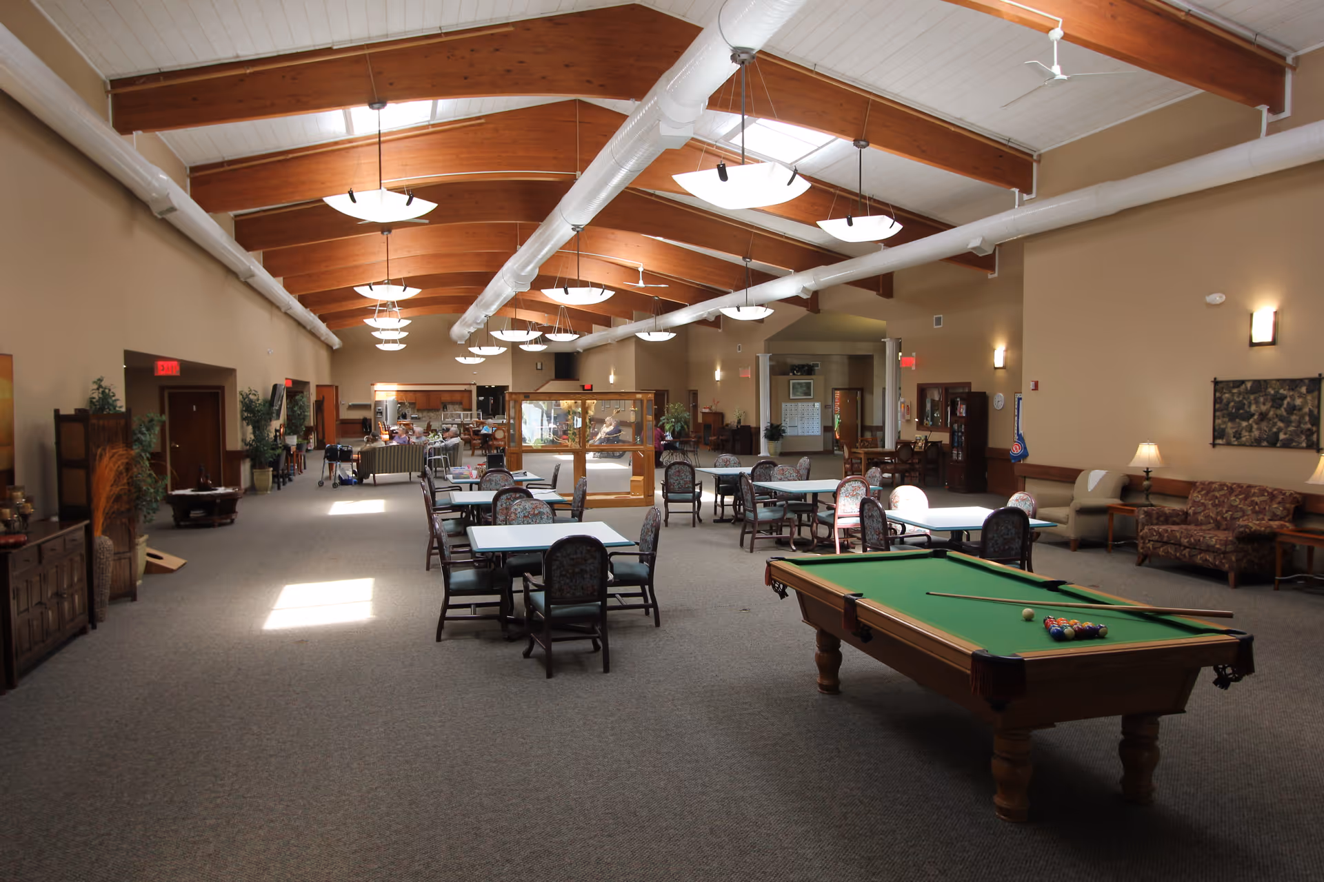 A spacious community room with high wooden beam ceilings and multiple hanging light fixtures. The room features several tables with chairs, a pool table with billiard balls and cues, and comfortable seating areas with sofas and armchairs. There are plants and decorative items along the walls, and a glass-enclosed area in the middle of the room. The space appears well-lit with natural light coming from windows and skylights.