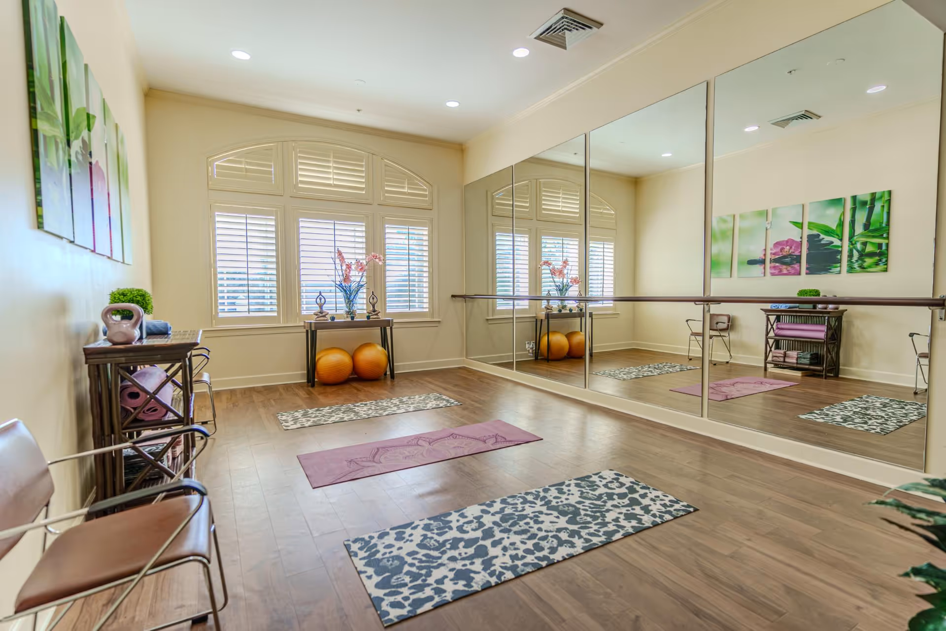 Bright exercise/yoga studio with a mirrored wall, several yoga mats on a wood floor, exercise balls, and a window with shutters.