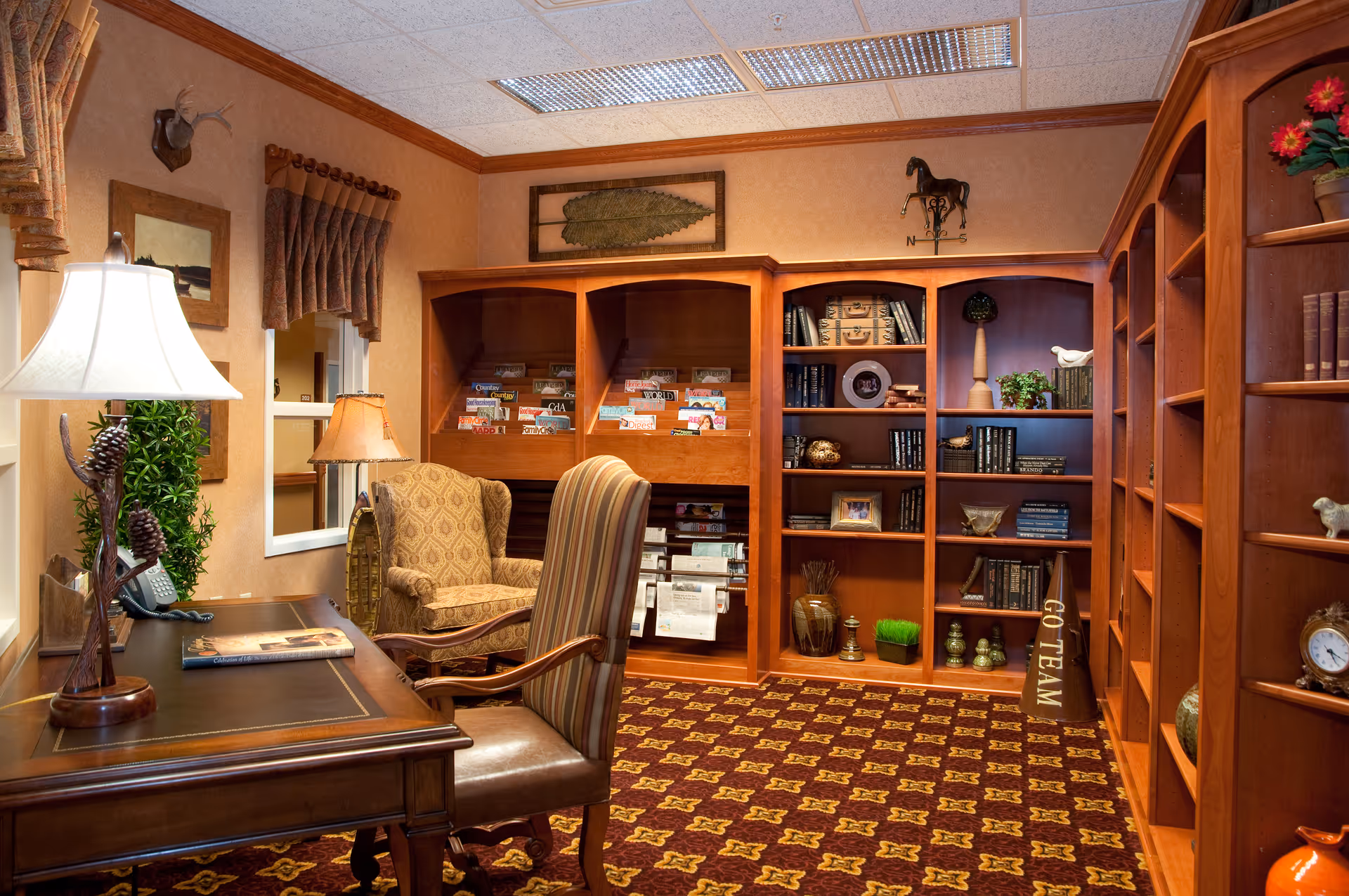 A cozy interior room with wooden bookshelves filled with books, decorative items, and magazines. There is a wooden desk with a lamp, a phone, and a book on it, accompanied by a striped upholstered chair. Another patterned armchair is placed near the shelves. The room has warm lighting, a patterned carpet, and wall decorations including a mounted deer head and framed artwork.