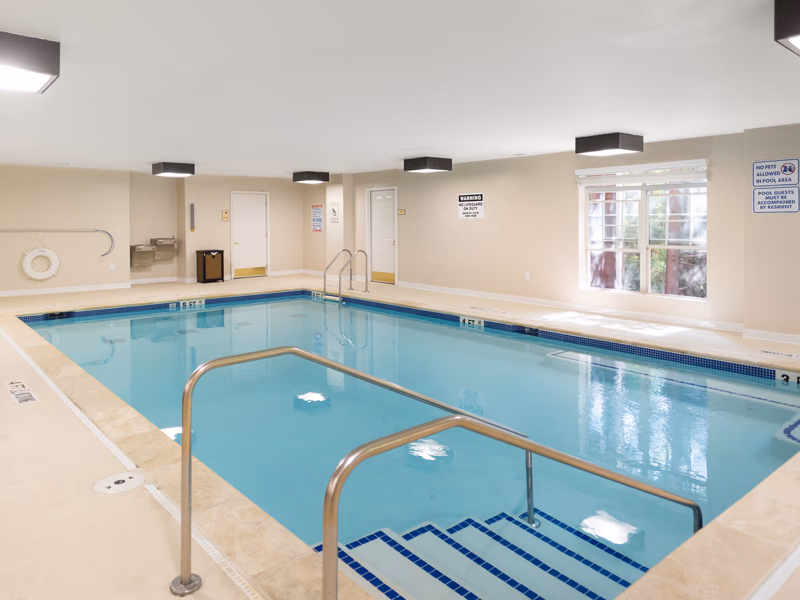 Indoor swimming pool with stainless steel handrails, tiled steps, windows, and safety signs on the walls.