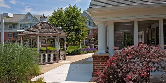 Outdoor view of a senior living facility featuring a paved walkway, a wooden gazebo, landscaped greenery including bushes and trees, and parts of the building with windows and a porch.