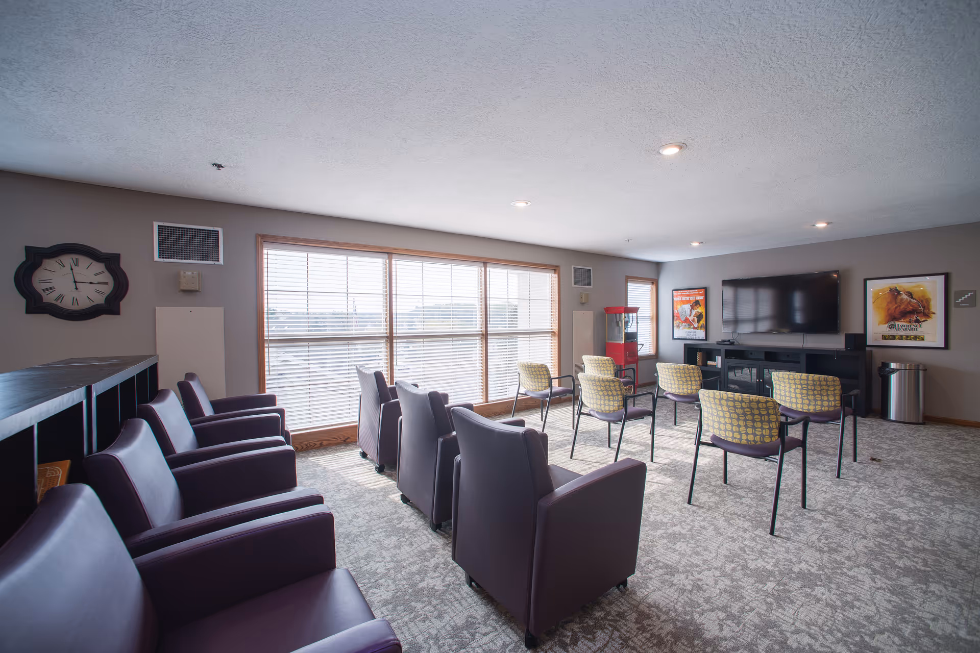A well-lit common room with multiple chairs arranged in rows facing a large flat-screen TV mounted on the wall. The room has large windows with blinds, a clock on the wall, framed posters, a popcorn machine, and a carpeted floor.