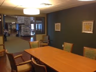 A conference or meeting room with a long wooden table surrounded by chairs. The room has a dark green accent wall with two framed artworks. Beyond the room, there is a view of a common area with a stone fireplace and additional seating.
