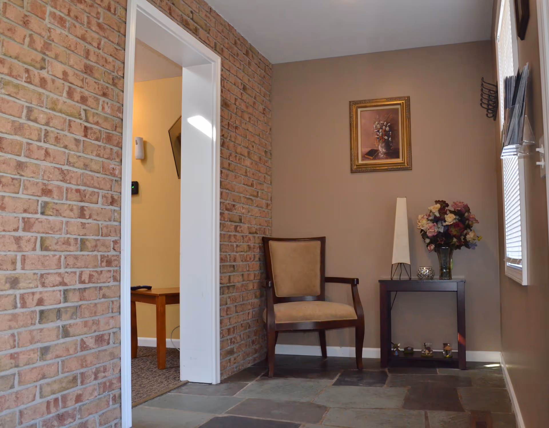 A small seating area with a wooden chair with beige upholstery next to a dark wooden side table holding a tall white lamp, a vase of colorful flowers, and a small decorative candle. The walls are painted beige, and one wall is covered with red brick. A framed painting of flowers hangs above the chair. There is a window with blinds on the right side, and an open doorway on the left leading to another room with a wooden table and carpeted floor.