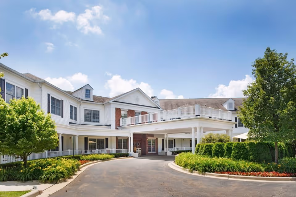 Exterior view of a senior living facility building with white siding and brick accents, surrounded by well-maintained landscaping including trees, bushes, and flowers under a blue sky with some clouds.