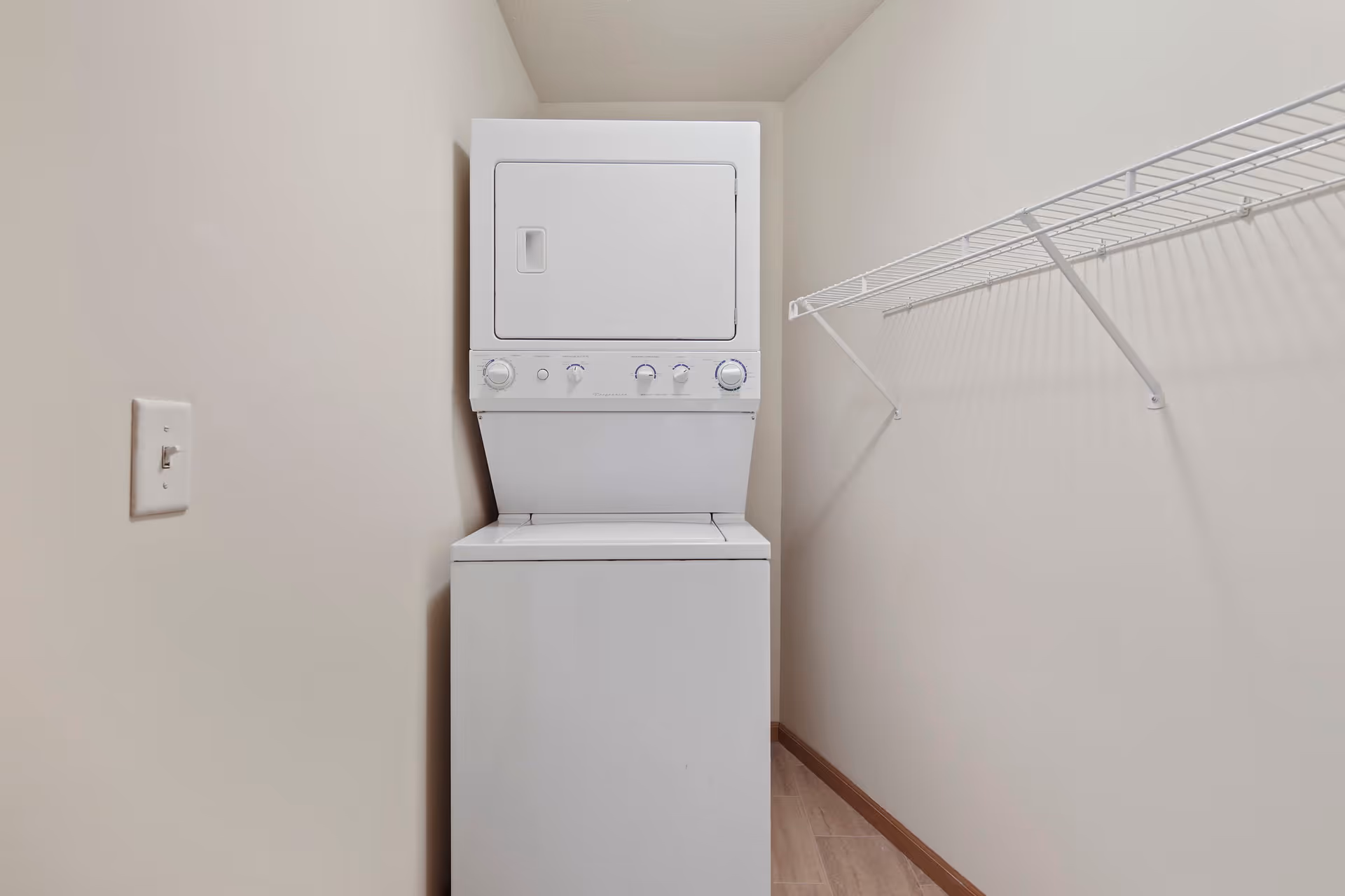 A small laundry area with a stacked white washer and dryer unit against a beige wall. To the right, there is a white wire shelf mounted on the wall. The floor is light-colored wood, and a light switch is visible on the left wall.
