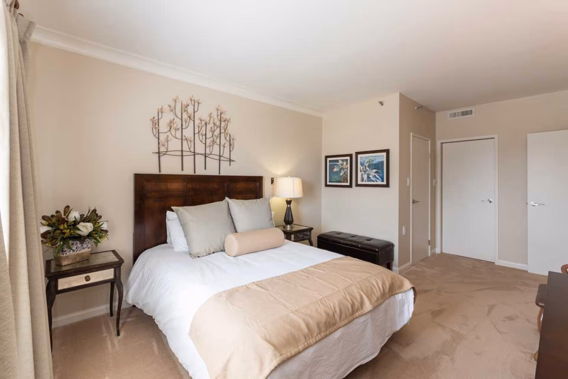 Bright, neutral-toned bedroom with a made bed, wooden headboard, nightstands, lamp and wall art.