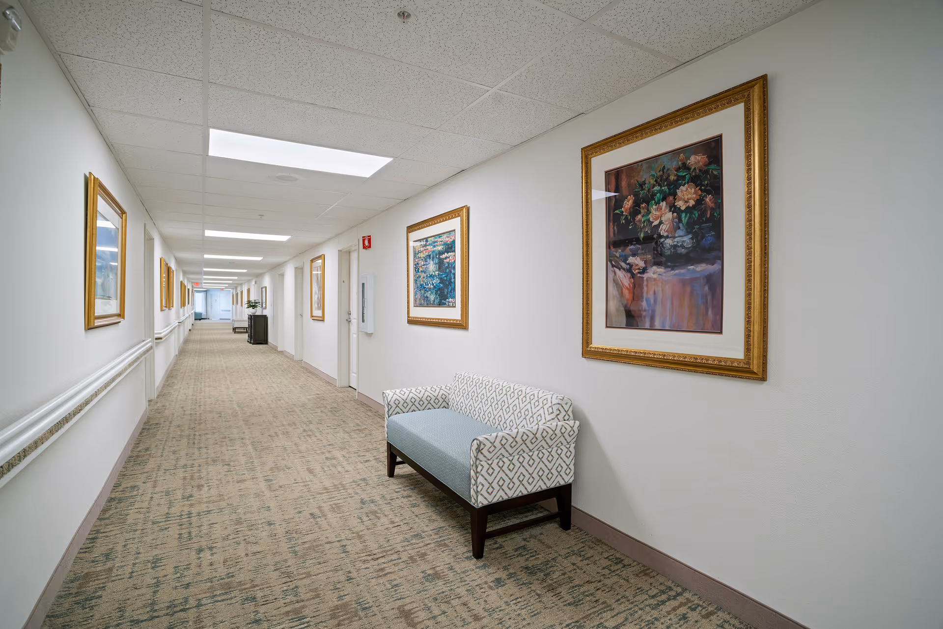 A long, well-lit hallway in a senior living facility with beige patterned carpet and white walls. The walls are decorated with framed paintings, and there is a patterned bench along the right side. Several doors line the hallway, and handrails run along both sides of the walls.