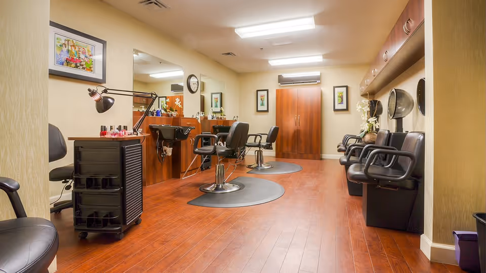 Interior view of a salon area in a senior living facility with wooden flooring, salon chairs, hair washing sinks, hair dryers, and a small cart with nail polish and a lamp. The walls are decorated with framed artwork and a clock.