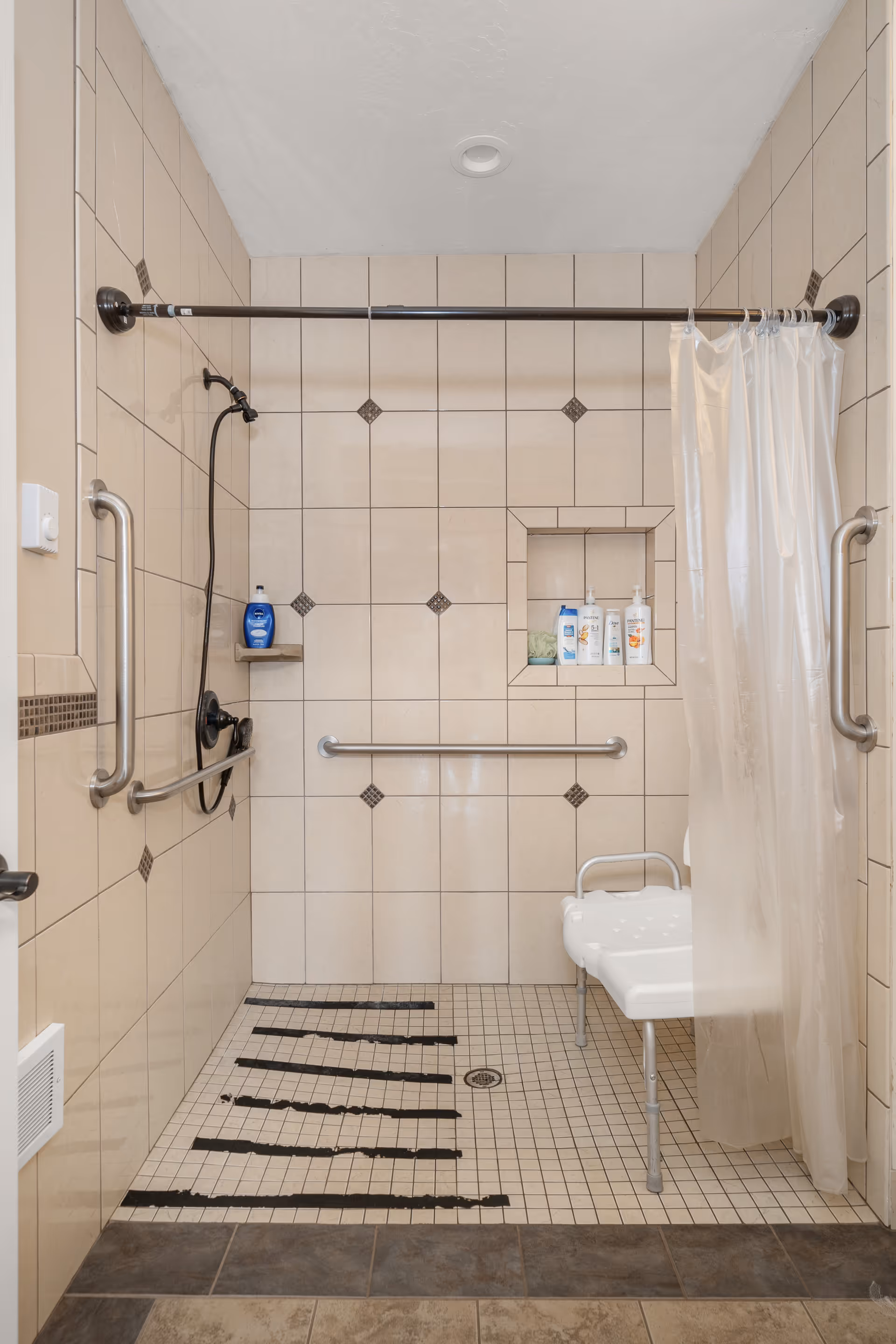 Tiled walk-in shower with grab bars, a shower seat, a handheld showerhead, and toiletries in a recessed shelf.