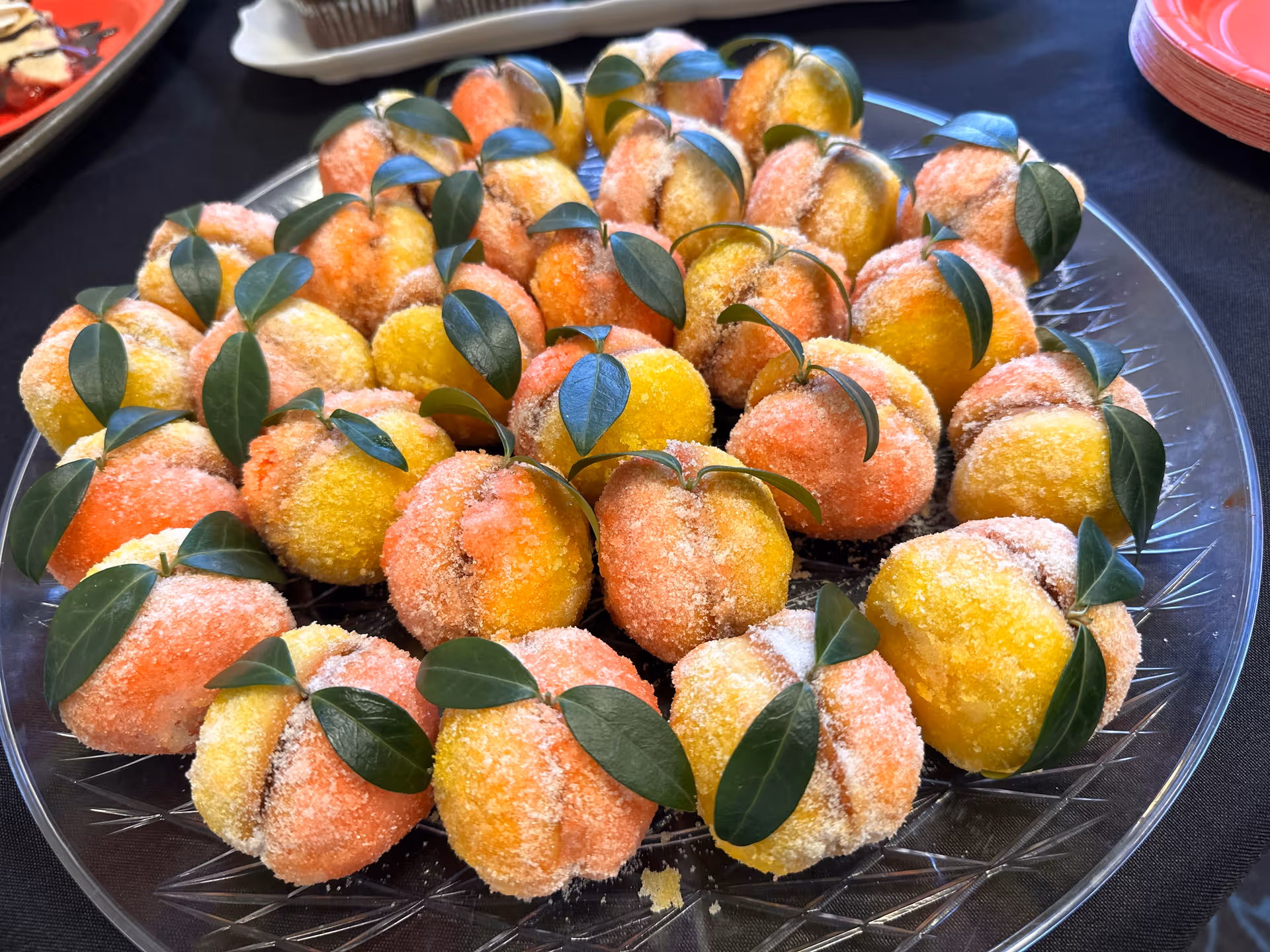 A clear glass plate filled with peach-shaped cookies dusted with sugar and decorated with small green leaves, arranged neatly on a black tablecloth.