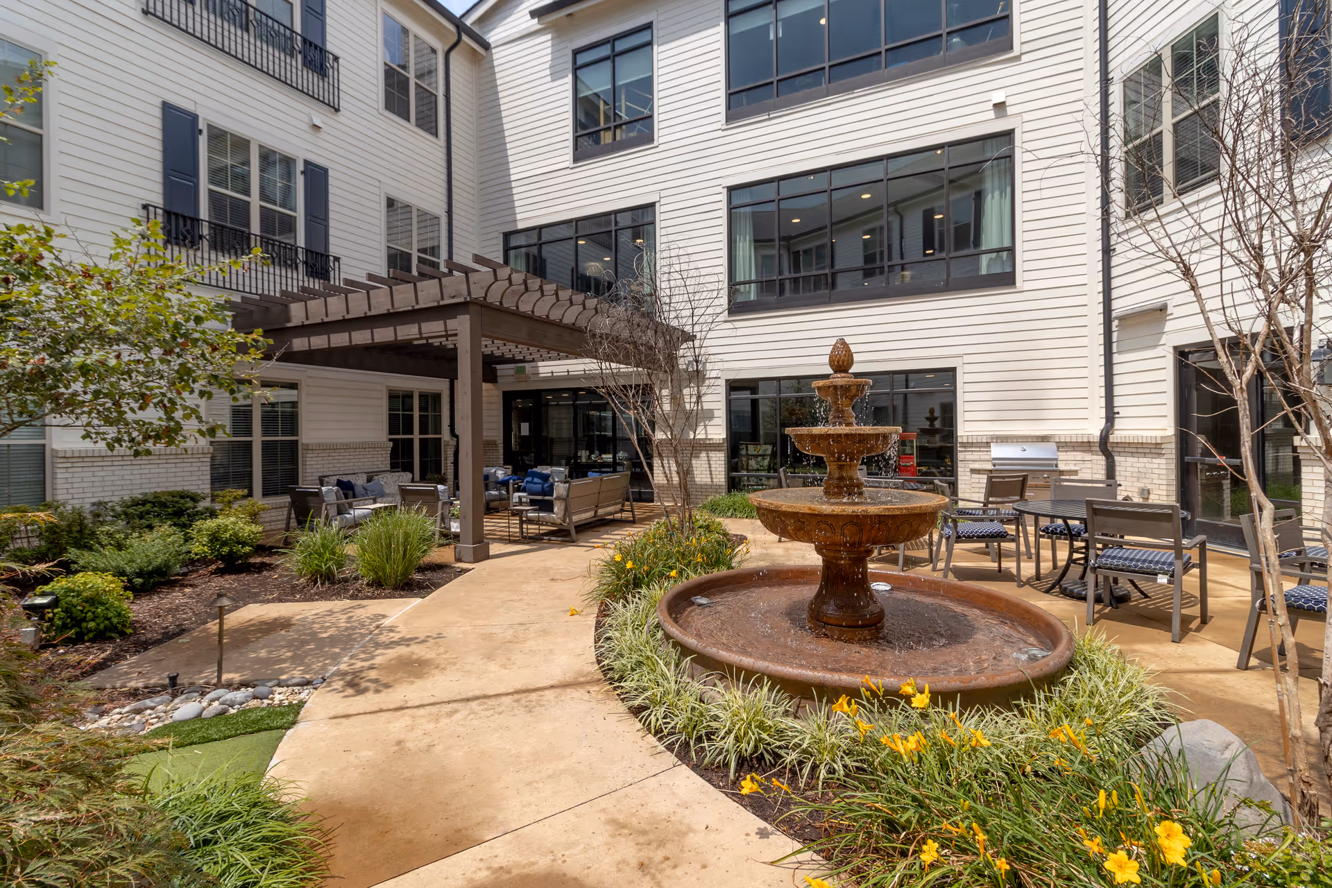 Outdoor courtyard with a tiered fountain, pergola-covered seating area, patio tables and landscaping in front of a multi-story building.