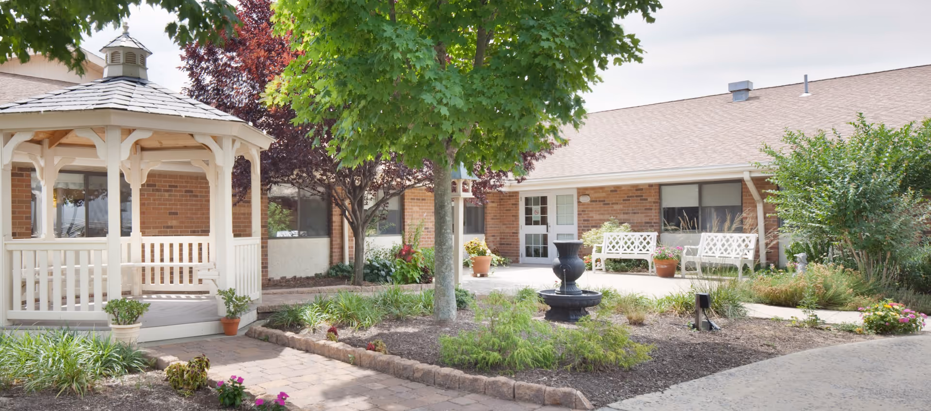 Courtyard with a white gazebo, central tree and fountain, benches and landscaping in front of a brick senior living building.