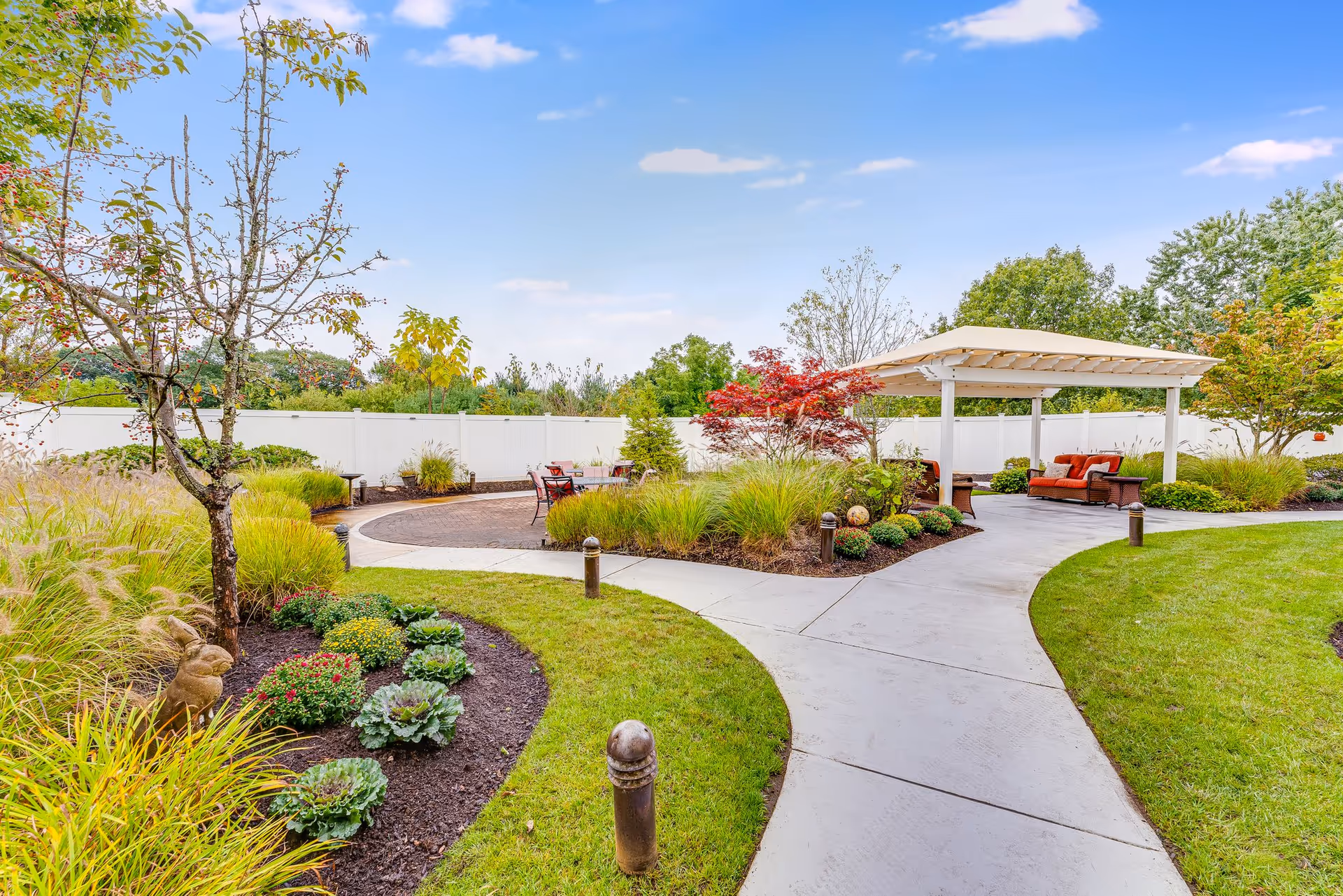 A well-maintained outdoor garden area with a curved concrete pathway leading to a covered seating area with cushioned chairs and a sofa. The garden features various plants, shrubs, and small trees, including a red-leafed tree, surrounded by a white fence under a blue sky with a few clouds.