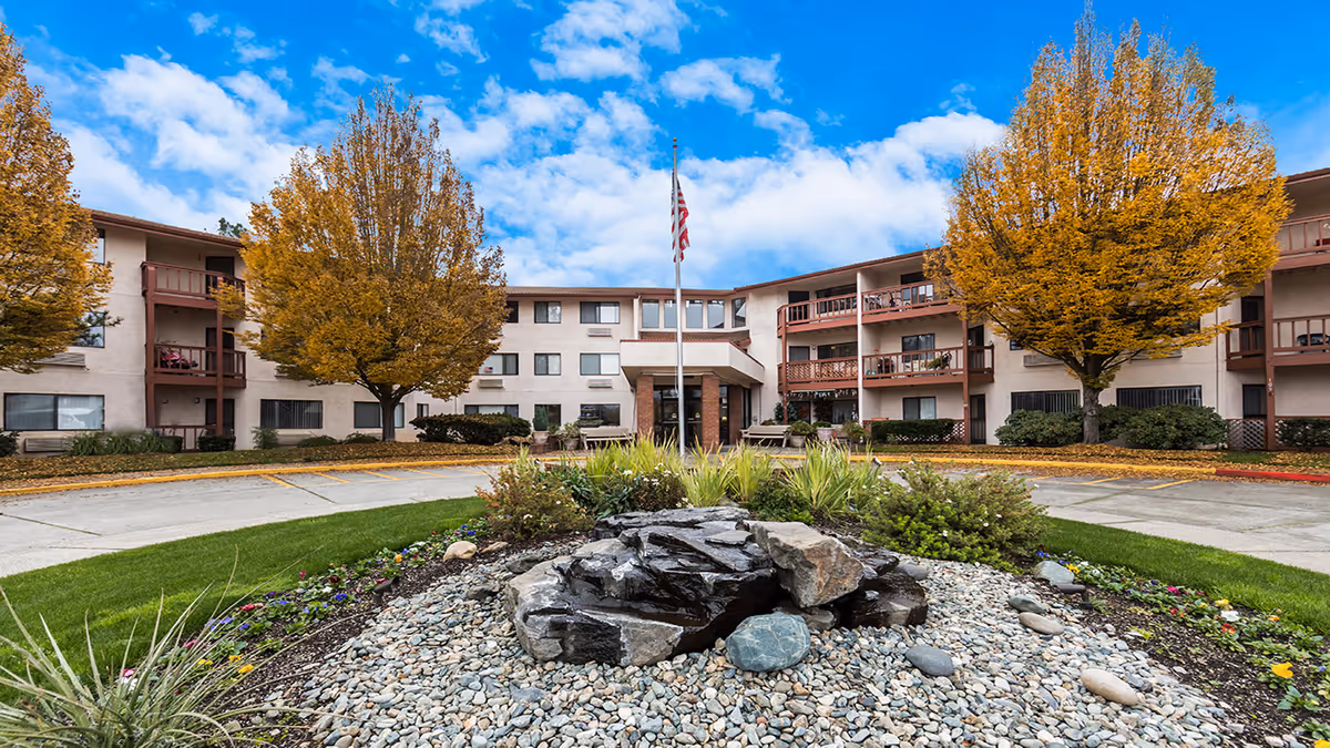 Exterior view of Solista Grants Pass facility with a circular driveway, landscaped garden with rocks and plants in the foreground, two large trees with autumn foliage, and a three-story building with balconies and windows under a partly cloudy blue sky.