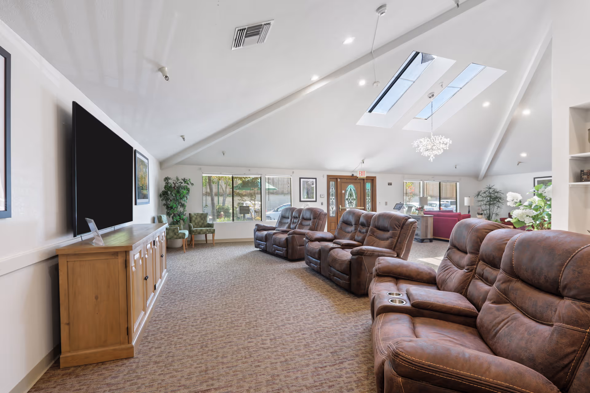 A spacious and well-lit assisted living facility common area with brown leather recliner chairs arranged in rows facing a large flat-screen TV mounted on the wall. The room features a high vaulted ceiling with skylights, carpeted floor, large windows letting in natural light, green plants, and a wooden cabinet under the TV. There is a wooden front door with glass panels and additional seating areas with chairs and a sofa in the background.