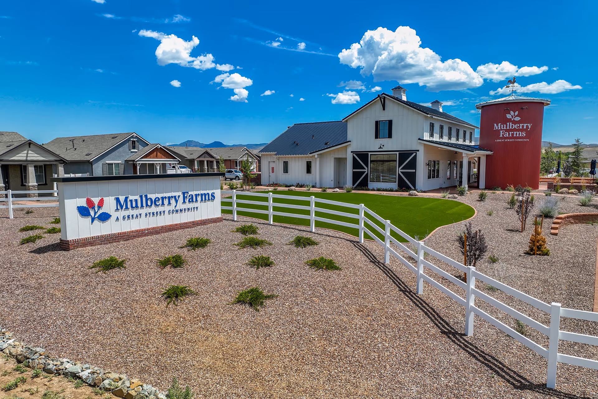 Exterior view of Mulberry Farms community with a white fence, landscaped gravel area with small plants, a green lawn, and a large white building with black trim and a red silo-like structure displaying the Mulberry Farms logo under a blue sky with clouds.