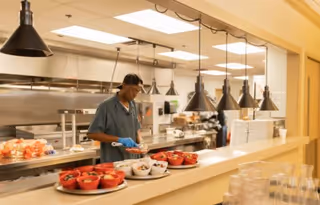 A staff member wearing gloves and a hairnet preparing food behind a serving counter in a kitchen area with multiple hanging pendant lights and trays of food bowls ready to be served.