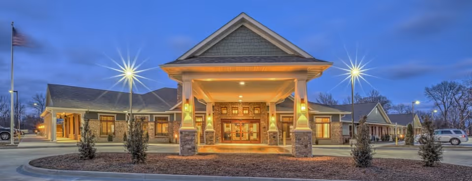 Exterior front view of a senior living facility building at dusk with warm lights illuminating the entrance. The building has a covered entryway supported by columns, a peaked roof, and surrounding parking areas with some cars and small trees.
