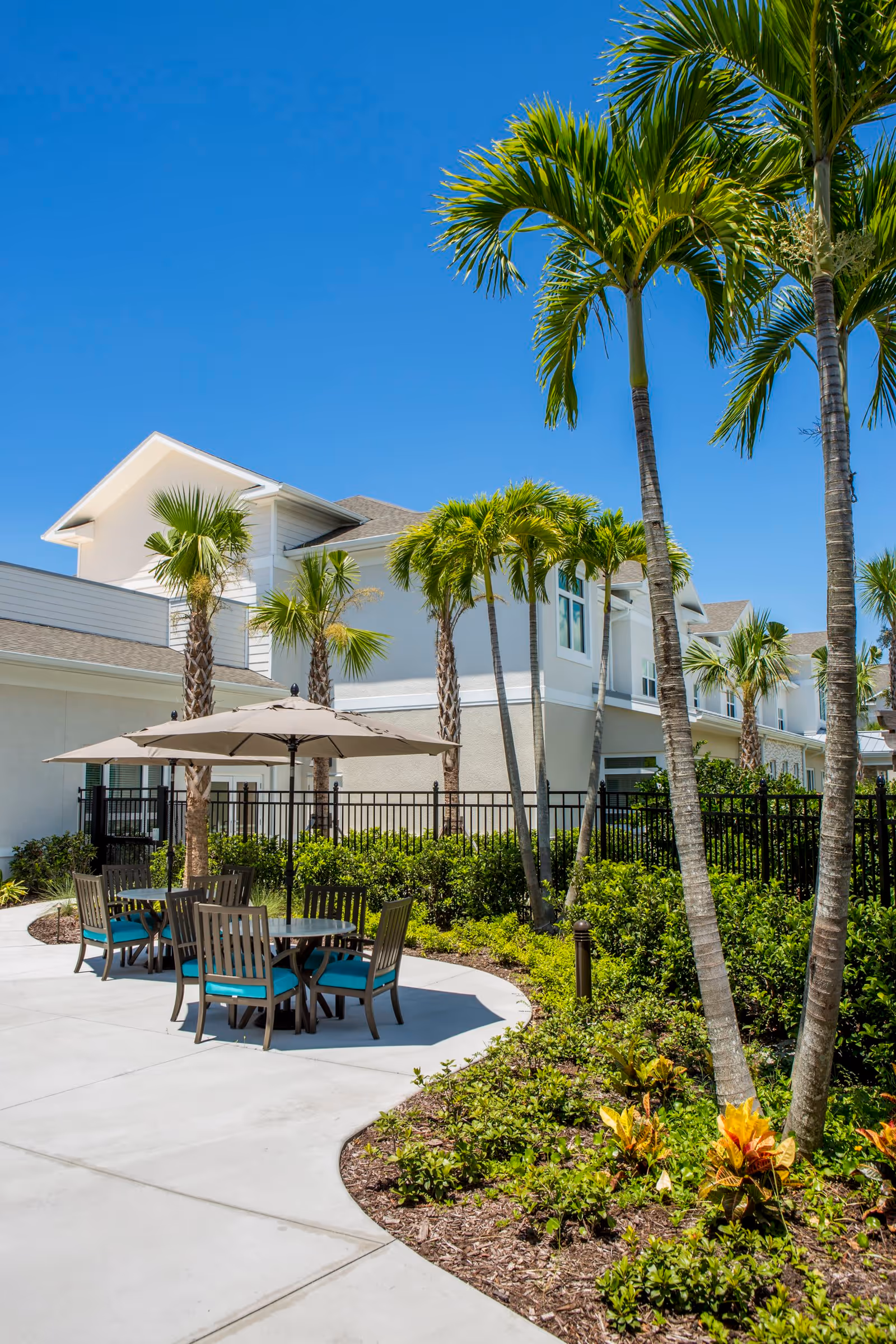 Outdoor patio area at Heritage Oaks of Englewood featuring round tables with umbrellas and chairs surrounded by palm trees and landscaped greenery under a clear blue sky.