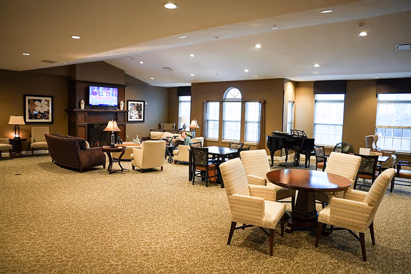 Spacious senior living common room with seating areas, round tables, a fireplace with TV, and large windows.
