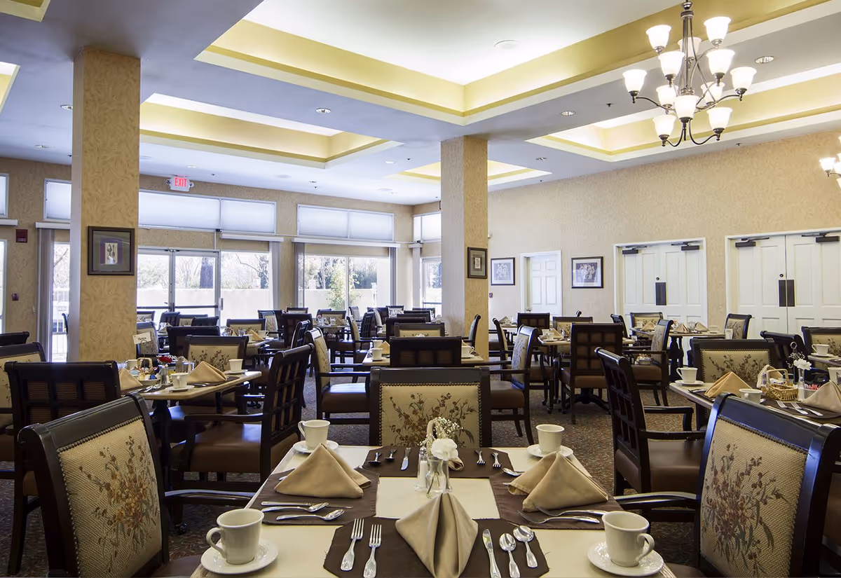 Bright, spacious dining room with multiple set tables, upholstered chairs, folded napkins, and chandeliers.