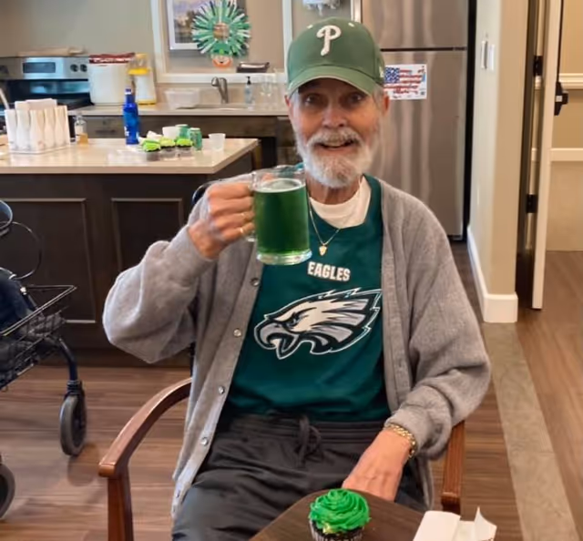 An elderly man wearing a Philadelphia Eagles shirt, gray cardigan, and green cap is sitting at a table in a kitchen or dining area. He is smiling and holding up a glass mug filled with a green beverage. On the table in front of him is a green-frosted cupcake. The background shows kitchen counters, a refrigerator, and some decorations.