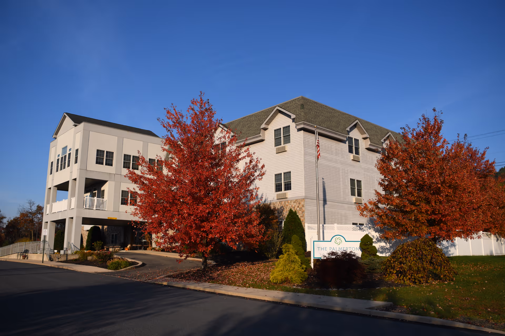 Front exterior of The Palmerton senior living building with red-leaved trees, a sign, and clear blue sky.